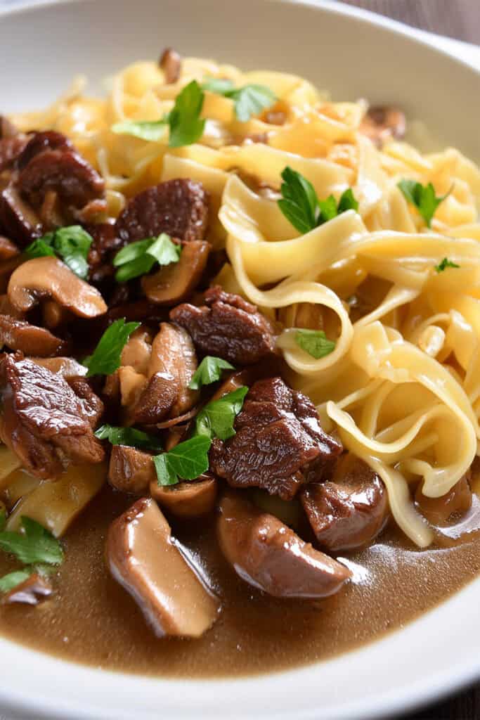 A plate of slow cooker beef with mushrooms stroganoff served with wide egg noodles, garnished with fresh parsley. The dish features tender chunks of beef and sliced mushrooms in a rich, savory sauce.