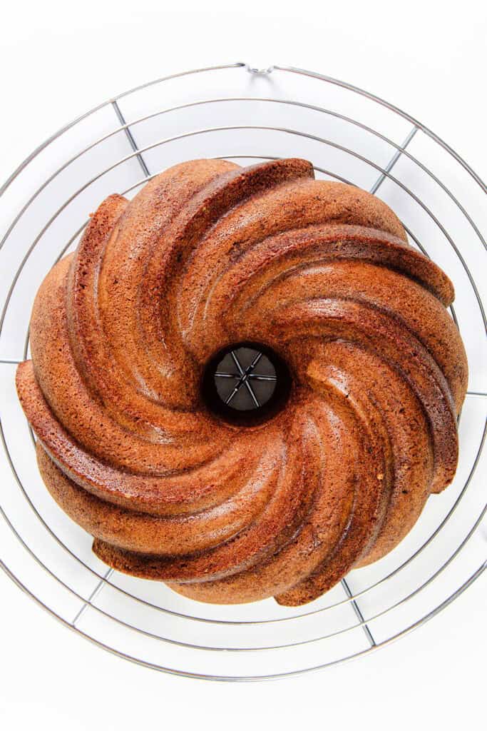 A top view of a decorative citrus and olive oil Bundt cake with a spiral pattern, placed on a round wire cooling rack against a white background. The cake has a golden-brown crust with a slight shine.