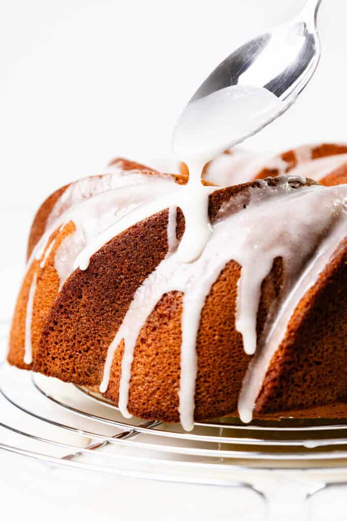 A close-up of a Citrus and Olive Oil Bundt Cake being drizzled with white icing from a spoon. The cake sits on a metal cooling rack, and the icing cascades down in thick streams. The white background ensures this delectable treat remains the focal point.