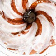 A close-up of a Citrus and Olive Oil Bundt cake with white icing drizzled over it, showcasing its spiral pattern. The cake rests on a cooling rack, allowing the icing to drip slightly through the gaps.