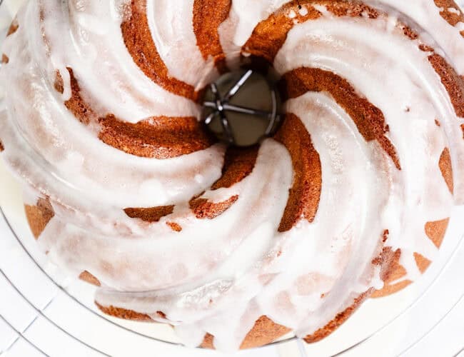 A close-up of a Citrus and Olive Oil Bundt cake with white icing drizzled over it, showcasing its spiral pattern. The cake rests on a cooling rack, allowing the icing to drip slightly through the gaps.