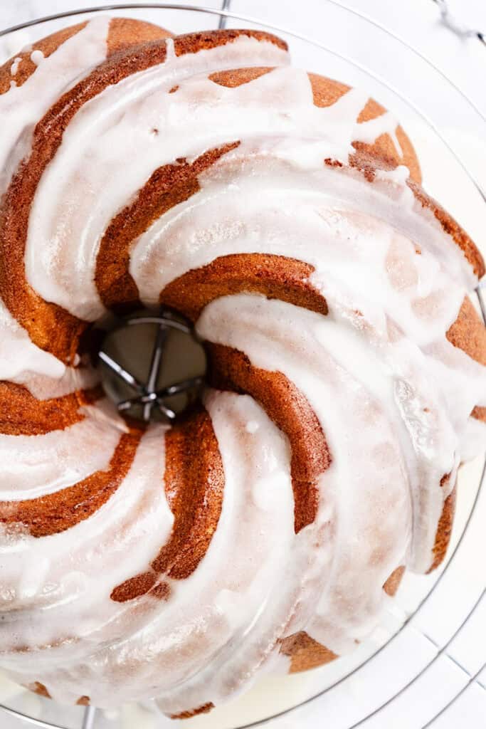 A Citrus and Olive Oil Bundt Cake rests on a wire cooling rack. The light brown cake is adorned with swirls of white icing drizzled over the top, creating a marbled effect as it sits elegantly on the marble countertop.