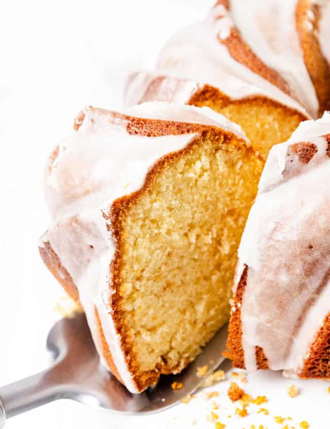 Close-up of a sliced Citrus and Olive Oil Bundt Cake with a golden-brown crust and white icing glaze. The cake is presented on a metal spatula against a white background.