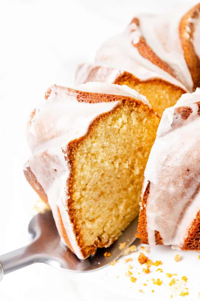 Close-up of a sliced Citrus and Olive Oil Bundt Cake with a golden-brown crust and white icing glaze. The cake is presented on a metal spatula against a white background.