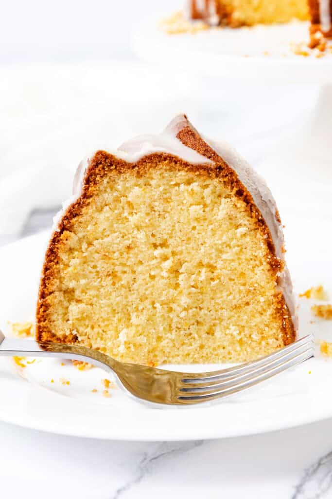 A slice of moist yellow cake with a light glaze on top, served on a white plate with a fork beside it. The cake, reminiscent of a citrus and olive oil Bundt creation, is placed on a marble surface, while the background features a blurred view of another cake on a stand.