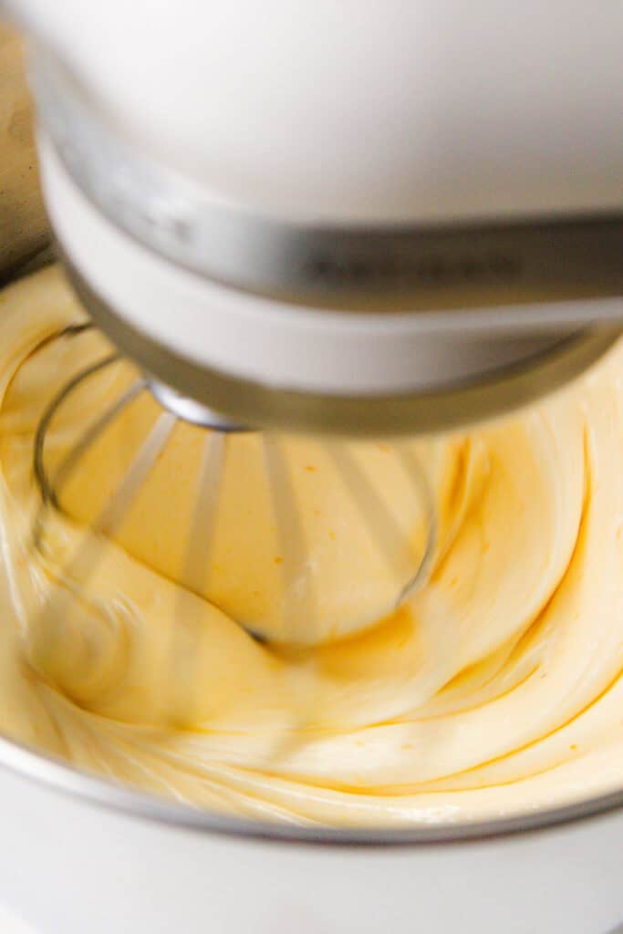 Close-up of a stand mixer with a wire whisk attachment blending a creamy, pale yellow batter in a metal mixing bowl. The citrus essence wafts as it whirls, while hints of olive oil promise richness to this Bundt cake masterpiece. Swirls dance elegantly in the smooth mixture.