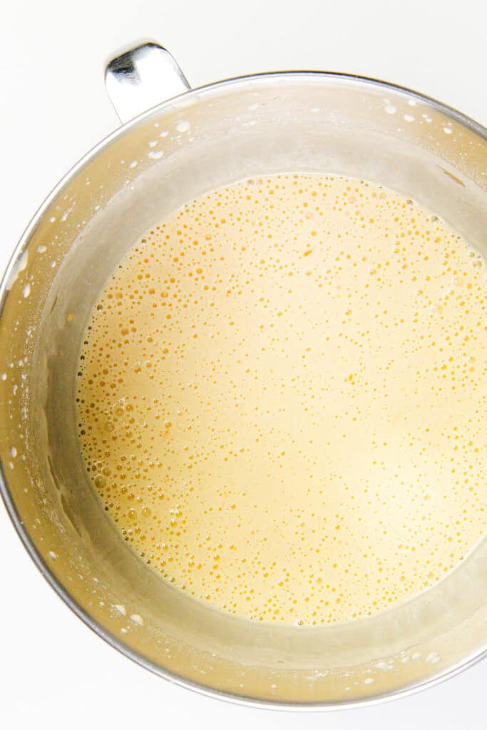 A metal mixing bowl filled with a frothy, light beige batter for a citrus and olive oil bundt cake is shown from above. The surface has tiny bubbles, and there is a handle on the left side of the bowl. The background is plain white.