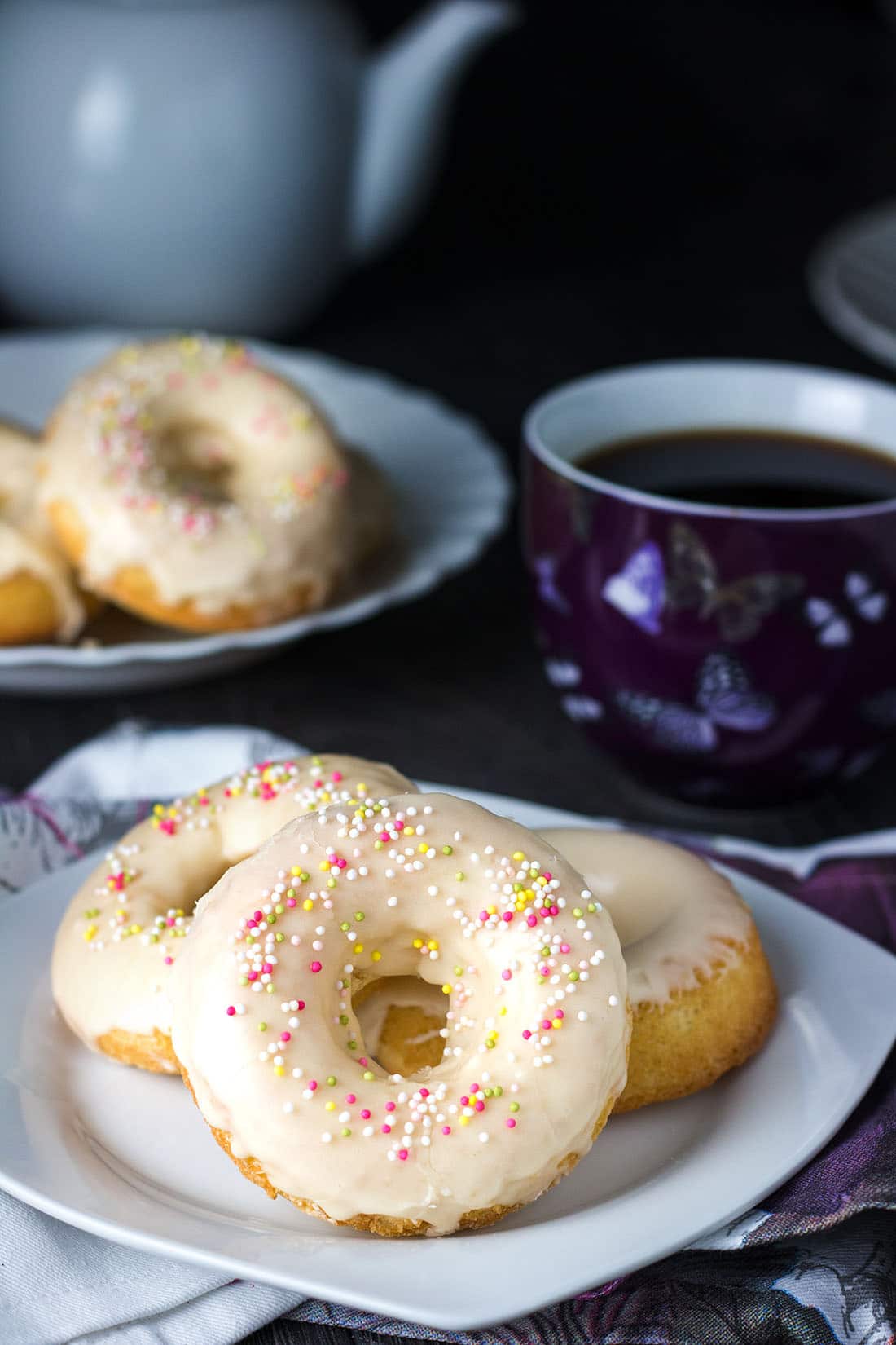 Easy Baked Iced Vanilla Doughnuts on a plate with the rest on a platter in the background an a mug full of coffee next to the plate.