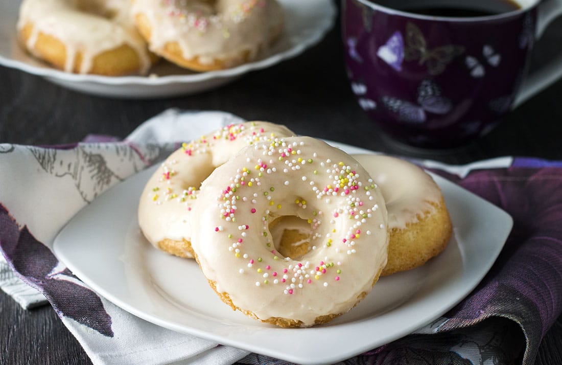 A plate with three iced vanilla doughnuts with colored sprinkles and a mug of coffee in the background.