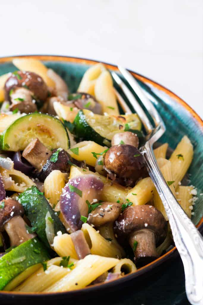 A bowl of roasted vegetable pasta with penne, sautéed zucchini, whole mushrooms, and red onions, garnished with chopped parsley and grated cheese, with a fork on the side.