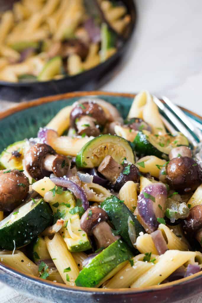 A bowl of roasted vegetable pasta featuring penne, sliced zucchini, brown mushrooms, and red onions, garnished with parsley and grated cheese, with a blurred serving dish in the background.