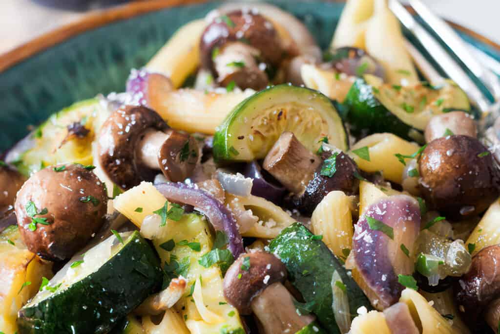 A close-up view of roasted vegetable pasta with zucchini, brown mushrooms, red onions, and parsley on penne, sprinkled with coarse salt and grated cheese.