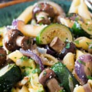 A close-up view of roasted vegetable pasta with zucchini, brown mushrooms, red onions, and parsley on penne, sprinkled with coarse salt and grated cheese.