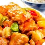 a close up of a bowl full of Sweet and sour chicken with white rice in the background