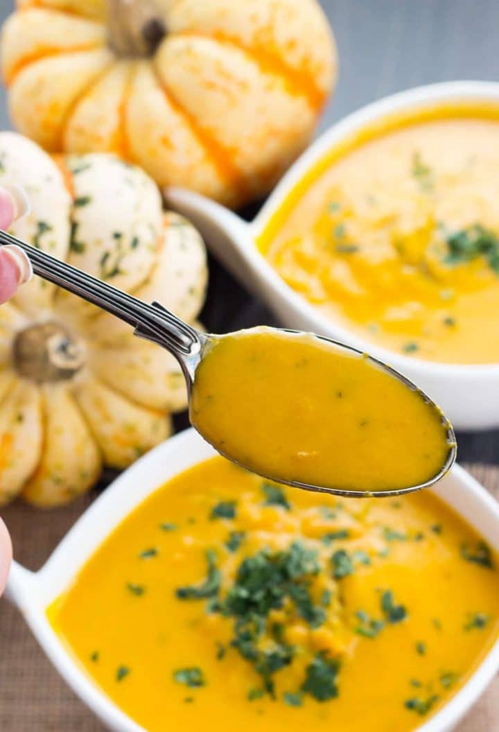 A spoonful of the winter squash soup over the bowl with whole winter squash in the background.