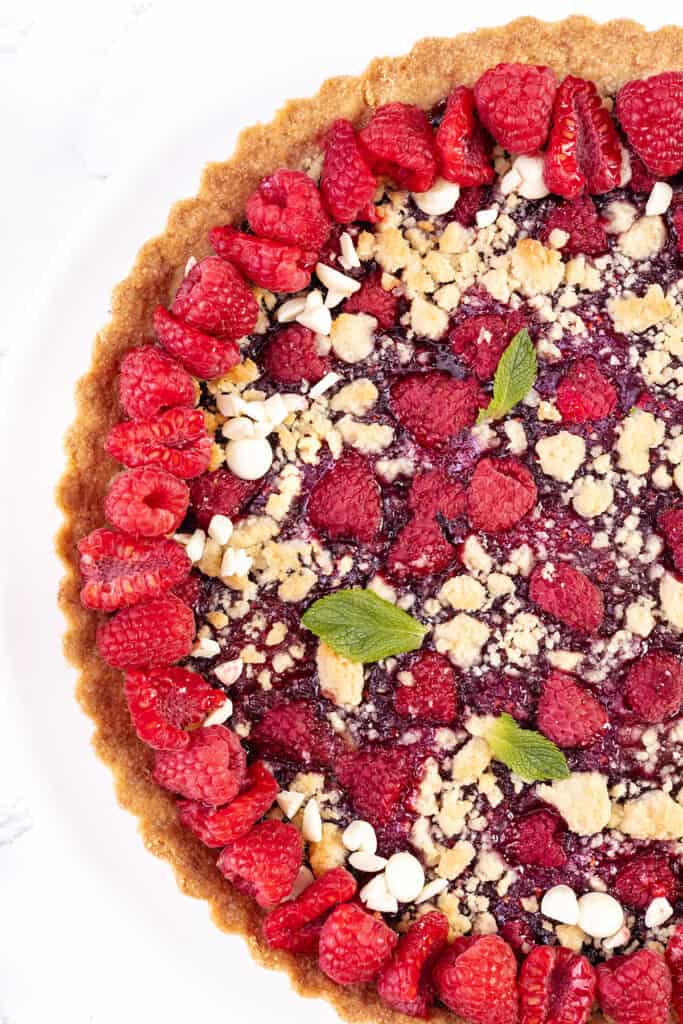 A close-up of a vibrant raspberry tart with a crumbly topping and garnished with fresh mint leaves. Whole raspberries adorn the edges, sprinkled with white chocolate chips. The golden brown crust contrasts beautifully with the vivid red filling.