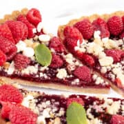 A close-up of a raspberry tart with a crumbly crust, topped with fresh raspberries, white chocolate chips, and a few mint leaves. A slice is being pulled out with a pie server, highlighting the juicy raspberry filling in this delightful raspberry tart.