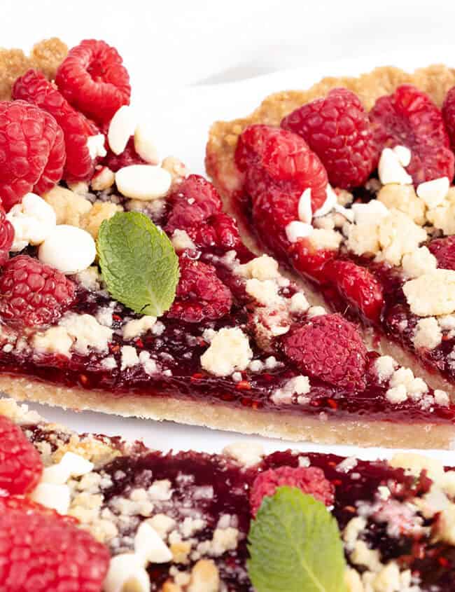 A close-up of a raspberry tart with a crumbly crust, topped with fresh raspberries, white chocolate chips, and a few mint leaves. A slice is being pulled out with a pie server, highlighting the juicy raspberry filling in this delightful raspberry tart.