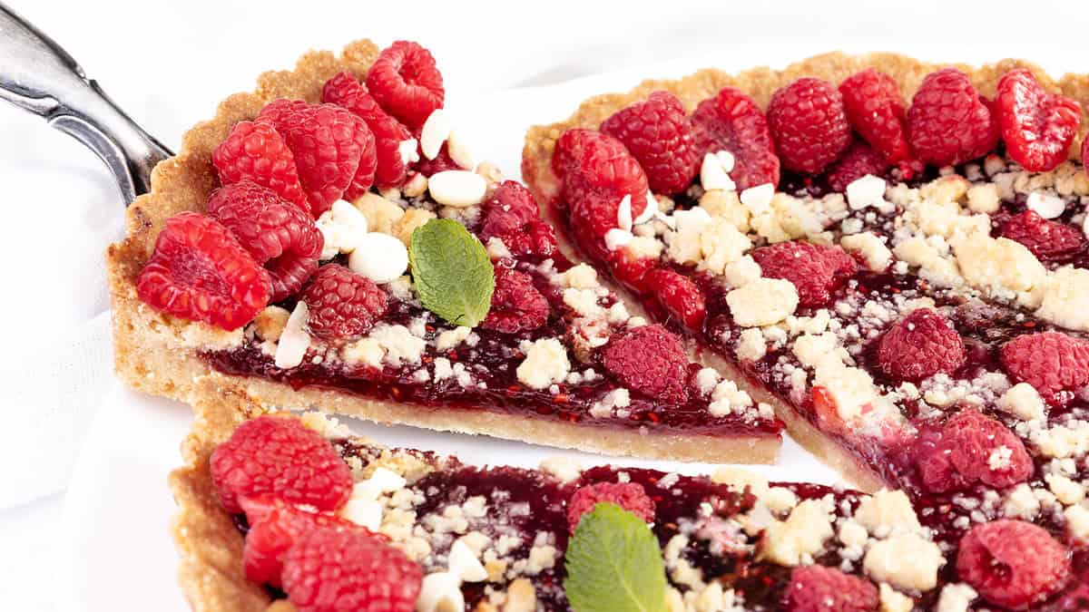 A close-up of a raspberry tart with a crumbly crust, topped with fresh raspberries, white chocolate chips, and a few mint leaves. A slice is being pulled out with a pie server, highlighting the juicy raspberry filling in this delightful raspberry tart.