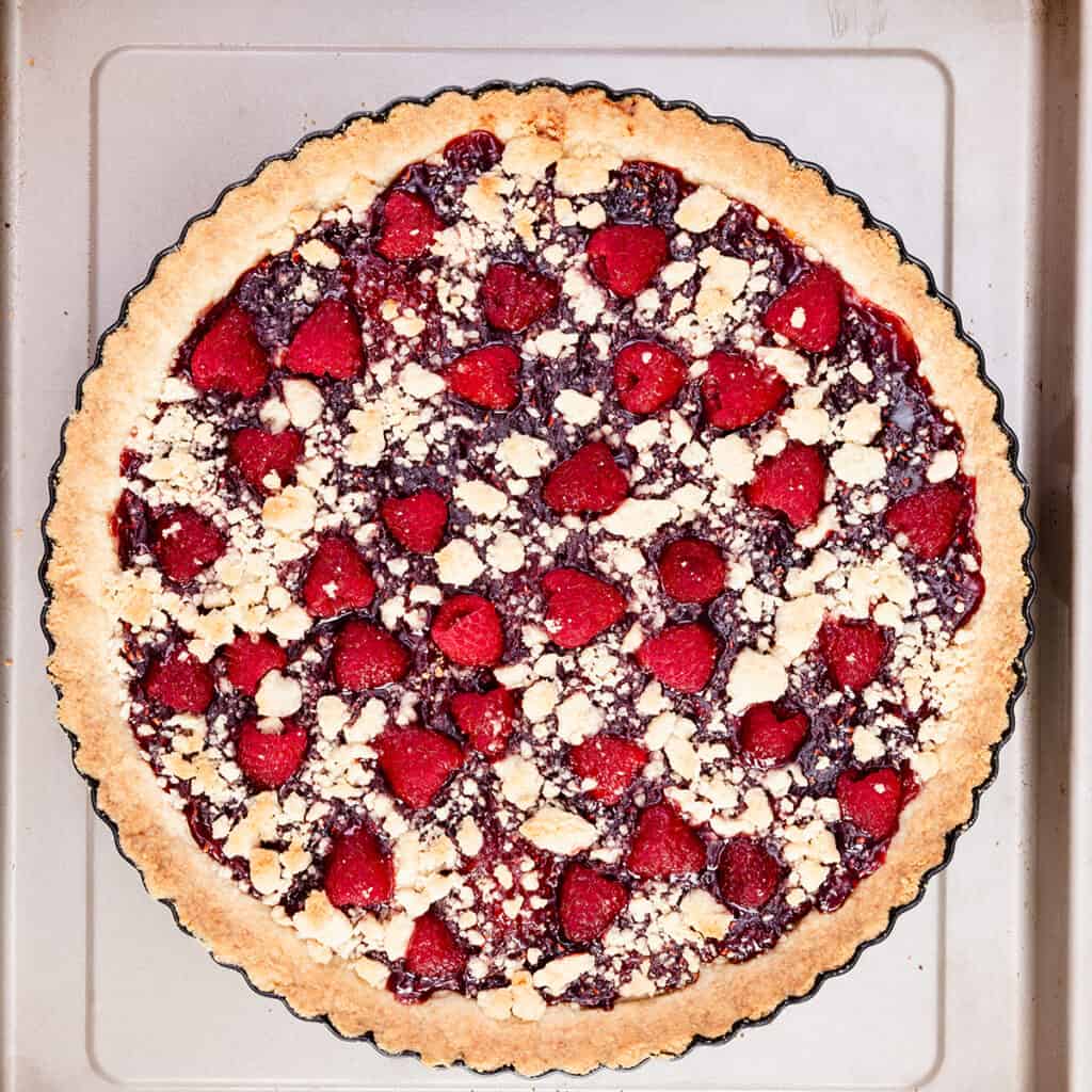 A close-up view of a freshly baked raspberry tart with a golden-brown crust. The raspberry tart is topped with vibrant red raspberries and sprinkled with crumbly streusel pieces, all set in a fluted tart pan on a baking sheet.