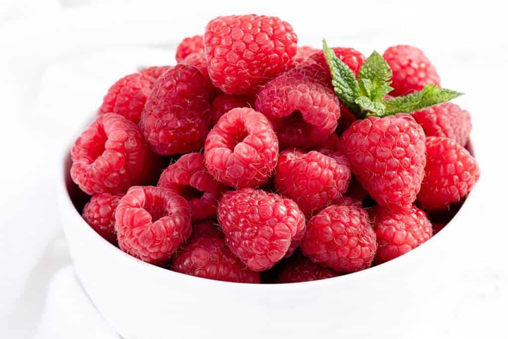 A white bowl is filled with fresh, ripe raspberries, featuring a vibrant red color. A small sprig of fresh green mint leaves rests on top, adding a touch of contrast. The background is soft and white, emphasizing the bright color of the raspberries.