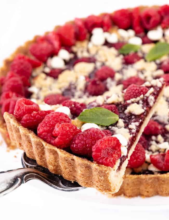 A close-up of a raspberry tart with a crumbly topping and white chocolate. The tart features a golden crust, and the raspberries are arranged neatly on top. A slice is being lifted out with a silver cake server. Mint leaves garnish the raspberry tart beautifully.