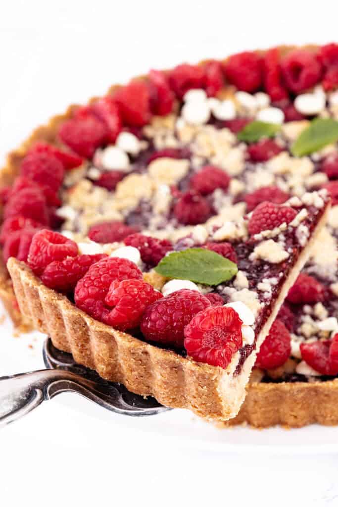 A close-up of a raspberry tart with a crumbly topping and white chocolate. The tart features a golden crust, and the raspberries are arranged neatly on top. A slice is being lifted out with a silver cake server. Mint leaves garnish the raspberry tart beautifully.