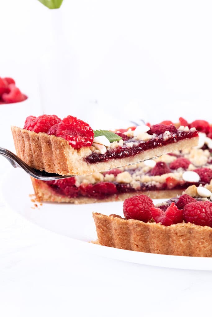 A slice of the delectable Raspberry Tart is being lifted from the whole tart with a serving spatula. The tart boasts a golden crust, a layer of raspberry filling, fresh raspberries on top, and is garnished with mint leaves and white almond slices. A white vase stands in the background.