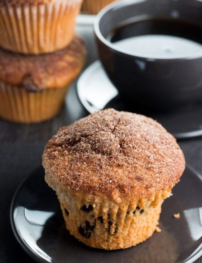 A Cinnamon Raisin Jumbo Muffin Topped with Cinnamon Sugar on a plate with a cup of coffee behind it.