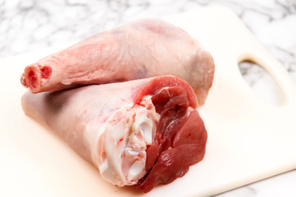 Two raw lamb shanks are placed on a white cutting board with a marble background. The shanks, perfect for slow-cooked lamb shanks, show both the bone and the red meat, ready for preparation in cooking.