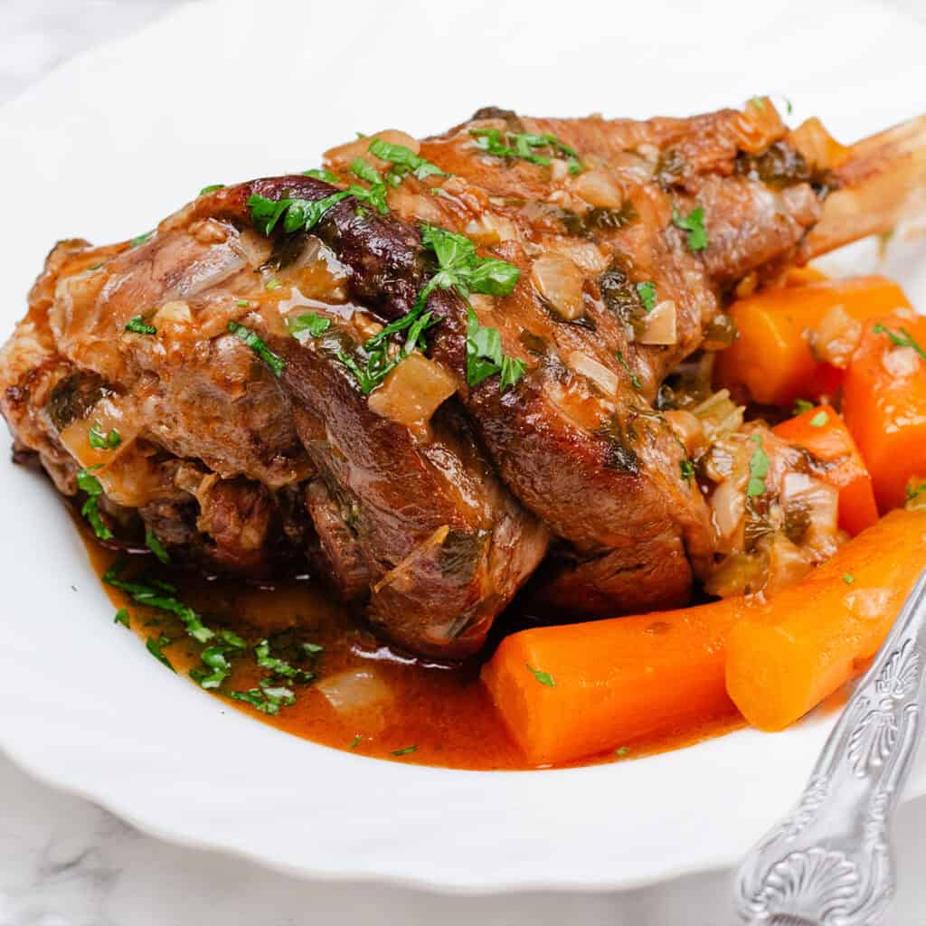 A slow-cooked lamb shank served on a white plate, accompanied by cooked carrot chunks. The lamb is topped with a rich gravy and garnished with chopped parsley. A silver fork rests on the plate's edge.