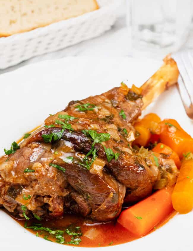 A close-up of a plate of slow cooked lamb shanks garnished with fresh parsley. The lamb shank is served with cooked carrots and herbs in a rich, brown sauce. Slices of bread are visible in a basket in the background.