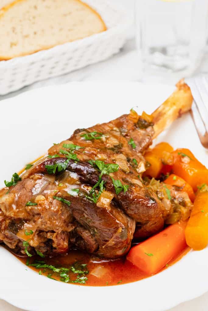 A close-up of a plate of slow cooked lamb shanks garnished with fresh parsley. The lamb shank is served with cooked carrots and herbs in a rich, brown sauce. Slices of bread are visible in a basket in the background.