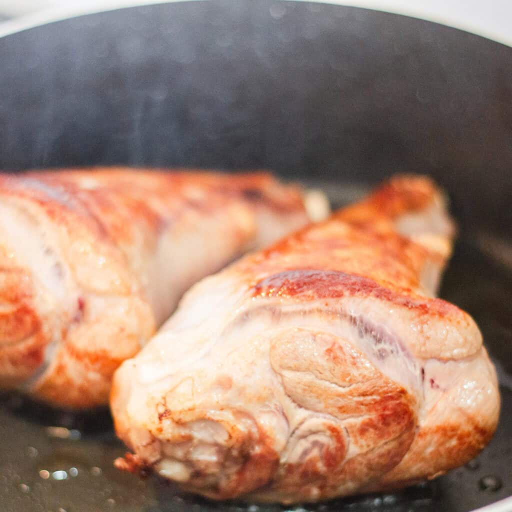 Close-up of two turkey legs being pan-fried in a black skillet. The turkey legs are golden brown, indicating they are in the process of being cooked. Light steam or smoke can be seen rising from the pan, reminiscent of slow-cooked lamb shanks simmering to perfection.