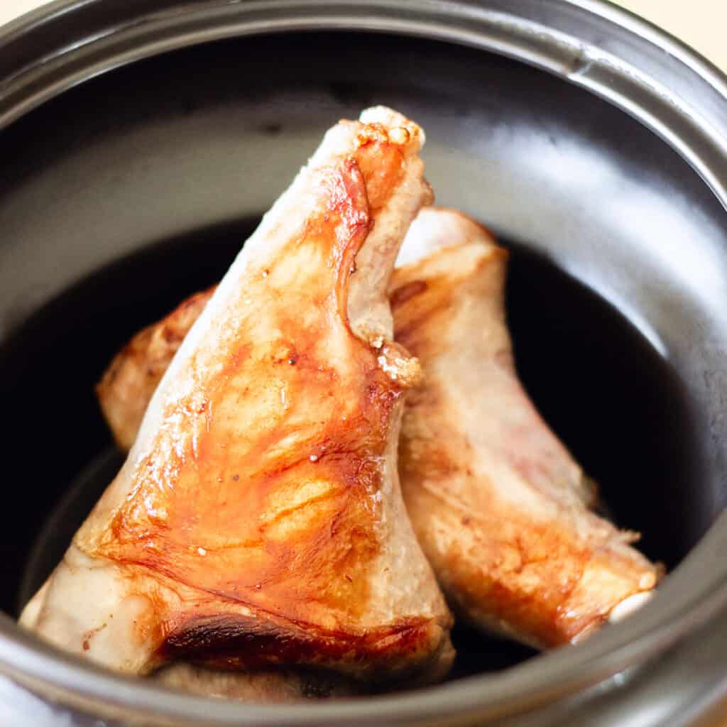 Close-up of two browned, slow cooked lamb shanks in a black slow cooker pot. The shanks are slightly tilted and have a crispy, golden outer layer, indicating they are well-cooked. The pot's interior contrasts with the rich color of the meat.