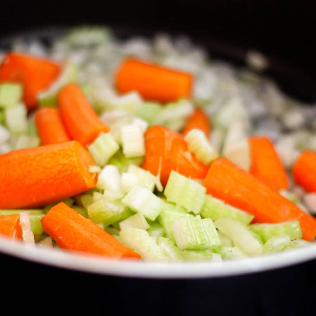 Close-up image of chopped carrots and celery being cooked in a pan. The bright orange of the carrot pieces contrasts with the green celery, creating a colorful and fresh vegetable medley. In the soft focus background, you can almost imagine slow cooked lamb shanks simmering away nearby.