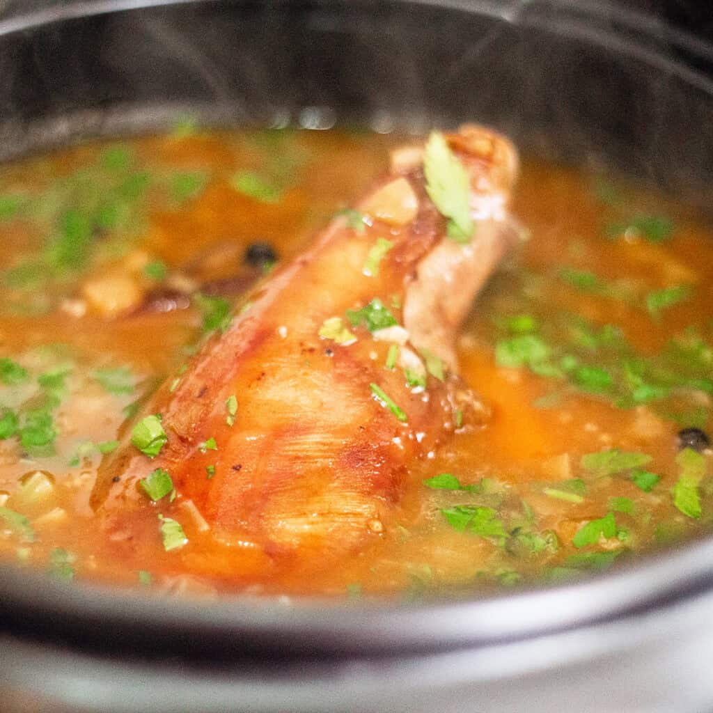 A close-up view of a steaming pot containing slow cooked lamb shanks submerged in a rich broth, garnished with fresh chopped herbs. The broth has a reddish tint and is thick with visible chunks of vegetables and seasonings.