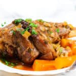 A plated dish featuring flavorful slow-cooked lamb shanks garnished with fresh parsley. The lamb is served with tender cooked carrots in a rich sauce, all presented on a white plate. A fork rests on the side of the plate, and a glass of water is visible in the background.