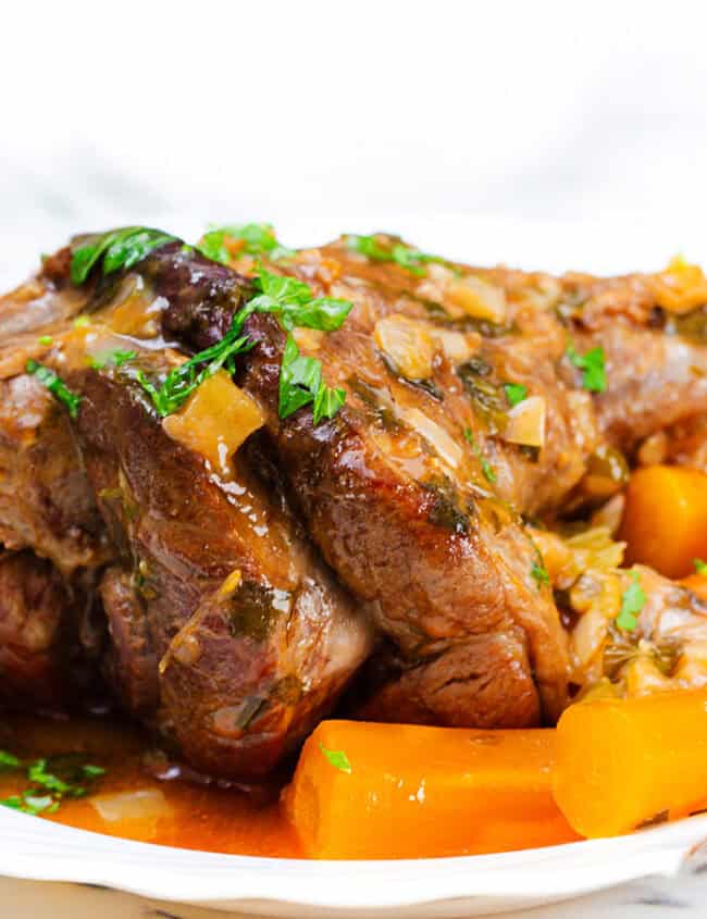 A plated dish featuring flavorful slow-cooked lamb shanks garnished with fresh parsley. The lamb is served with tender cooked carrots in a rich sauce, all presented on a white plate. A fork rests on the side of the plate, and a glass of water is visible in the background.