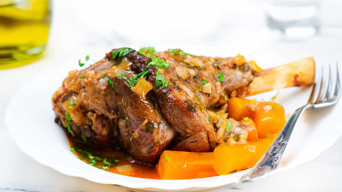 A plated dish featuring flavorful slow-cooked lamb shanks garnished with fresh parsley. The lamb is served with tender cooked carrots in a rich sauce, all presented on a white plate. A fork rests on the side of the plate, and a glass of water is visible in the background.