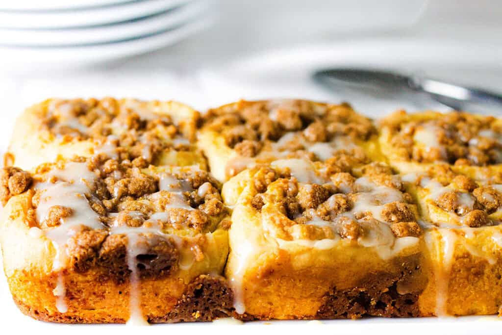 A close-up of six Cinnamon Crumb Buns topped with crumbly streusel and a glaze. The rolls are slightly golden brown, and the icing drips down the sides. White plates and a spoon are blurred in the background.