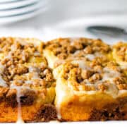 A close-up of six Cinnamon Crumb Buns topped with crumbly streusel and a glaze. The rolls are slightly golden brown, and the icing drips down the sides. White plates and a spoon are blurred in the background.