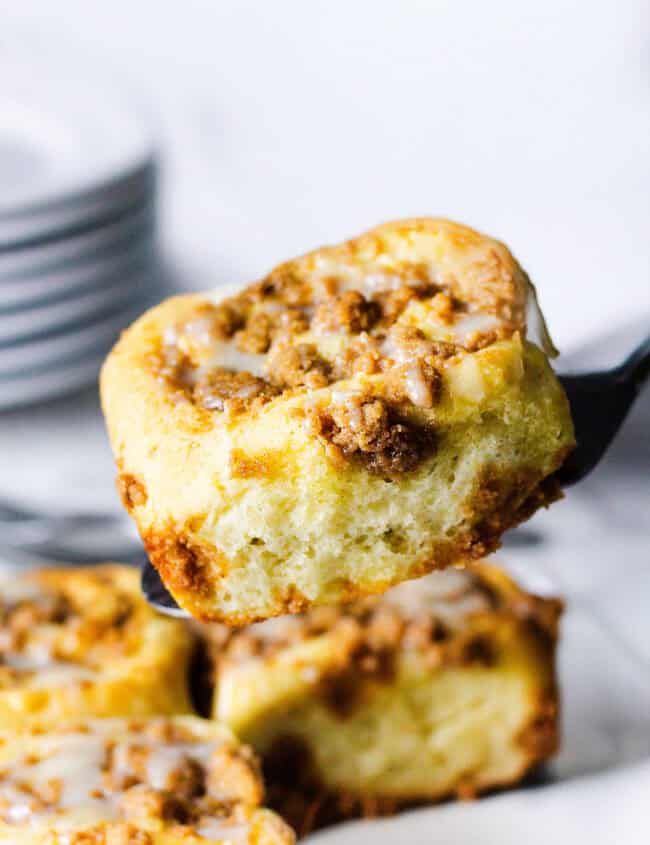 A close-up of a Cinnamon Crumb Bun being lifted by a spatula. The roll is topped with a crumble of brown sugar and nuts, and drizzled with a light glaze. In the background, a stack of white plates can be partially seen.