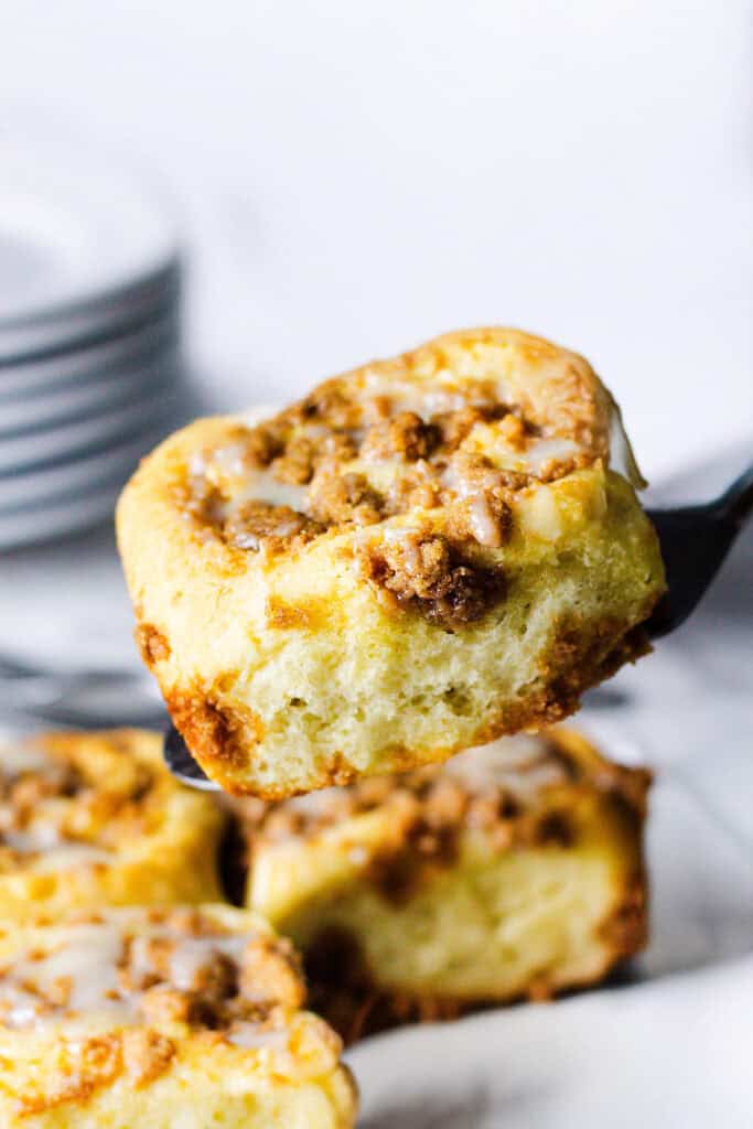 A close-up of a Cinnamon Crumb Bun being lifted by a spatula. The roll is topped with a crumble of brown sugar and nuts, and drizzled with a light glaze. In the background, a stack of white plates can be partially seen.