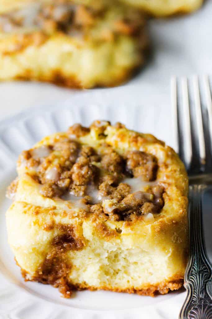 A close-up of a Cinnamon Crumb Bun on a white plate. The roll is topped with a crumbly streusel and drizzled with light icing. A fork lies next to the plate on the right side, partially visible. The background shows another blurred cinnamon roll.