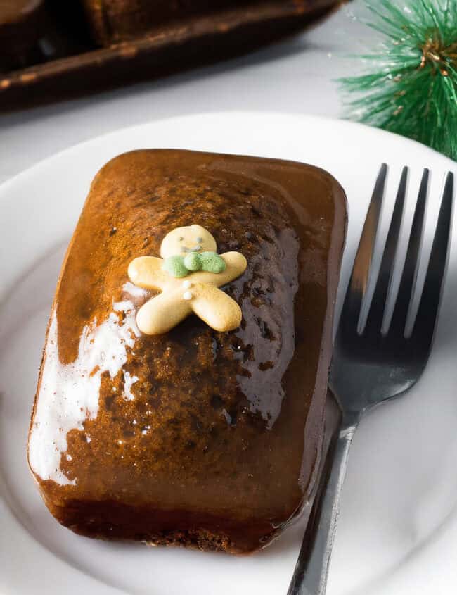 A glazed rectangular gingerbread cake sits on a white plate, crowned with a small gingerbread man decoration. Beside the cake, a fork awaits next to festive green decor, reminiscent of classic sticky toffee ginger cakes that evoke warm holiday cheer.