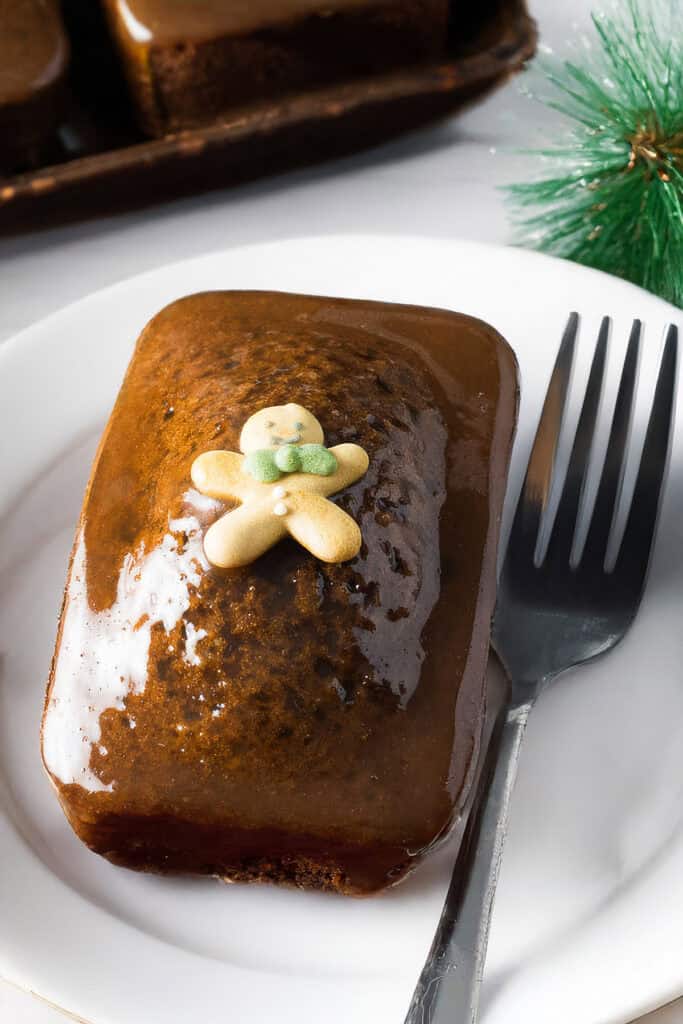 A glazed rectangular gingerbread cake sits on a white plate, crowned with a small gingerbread man decoration. Beside the cake, a fork awaits next to festive green decor, reminiscent of classic sticky toffee ginger cakes that evoke warm holiday cheer.