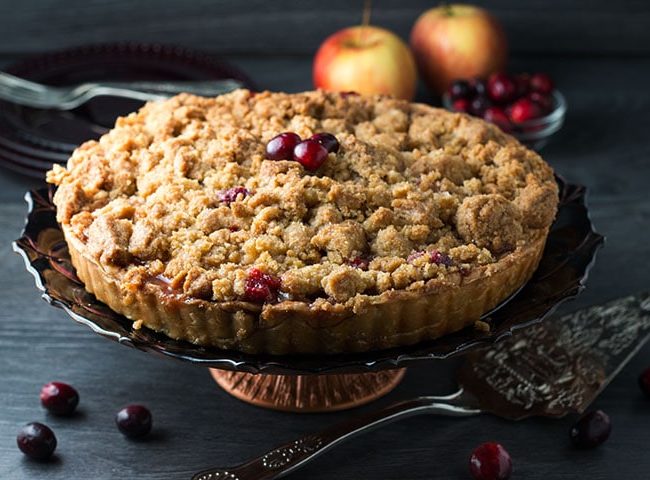 An apple cranberry crumb pie on a pie stand with cranberries on top, scattered on the table and some apples in the background.