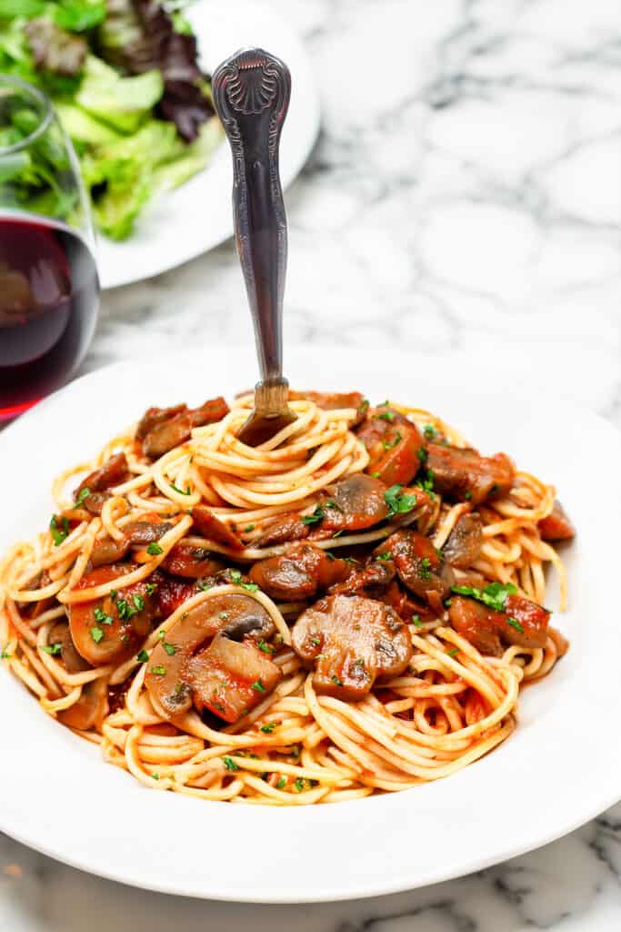 A round white plate overflowing with tangled spaghetti strands, coated in a rich red tomato sauce with earthy-colored sliced mushrooms throughout. Fresh green parsley flakes are sprinkled on top for garnish.