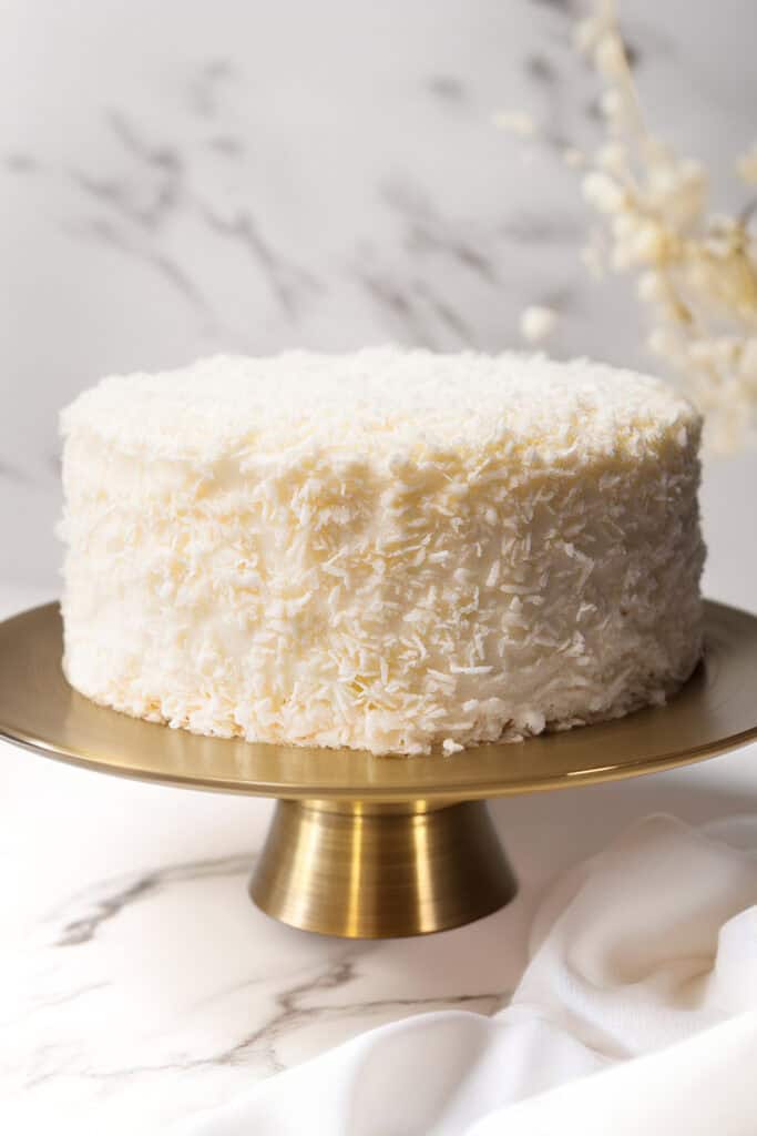 A round white Coconut Lovers Dream Cake with Coconut Buttercream Frosting is displayed on a golden cake stand. The background is softly blurred with white and gray tones, adding an elegant touch to the image. A piece of white fabric is draped to the side.